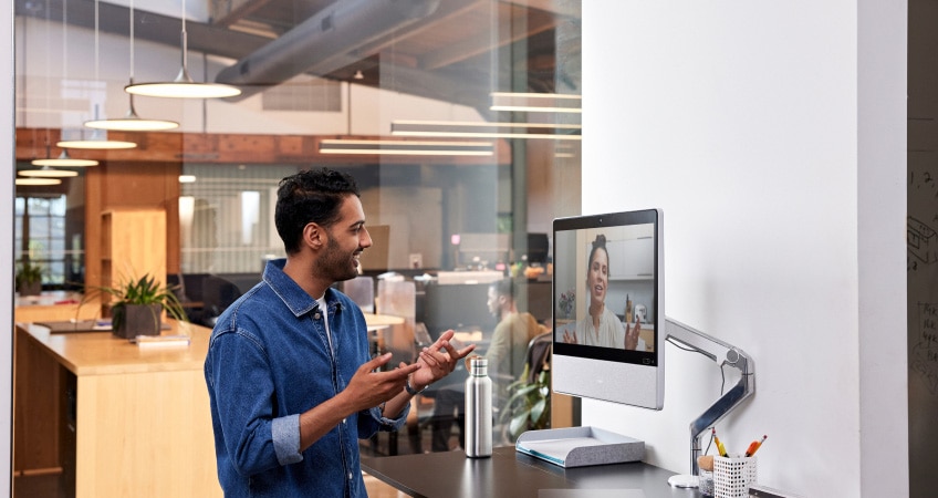  Person in a denim button down shirt at a standing desk in an office collaborates with a colleague via a video meeting.