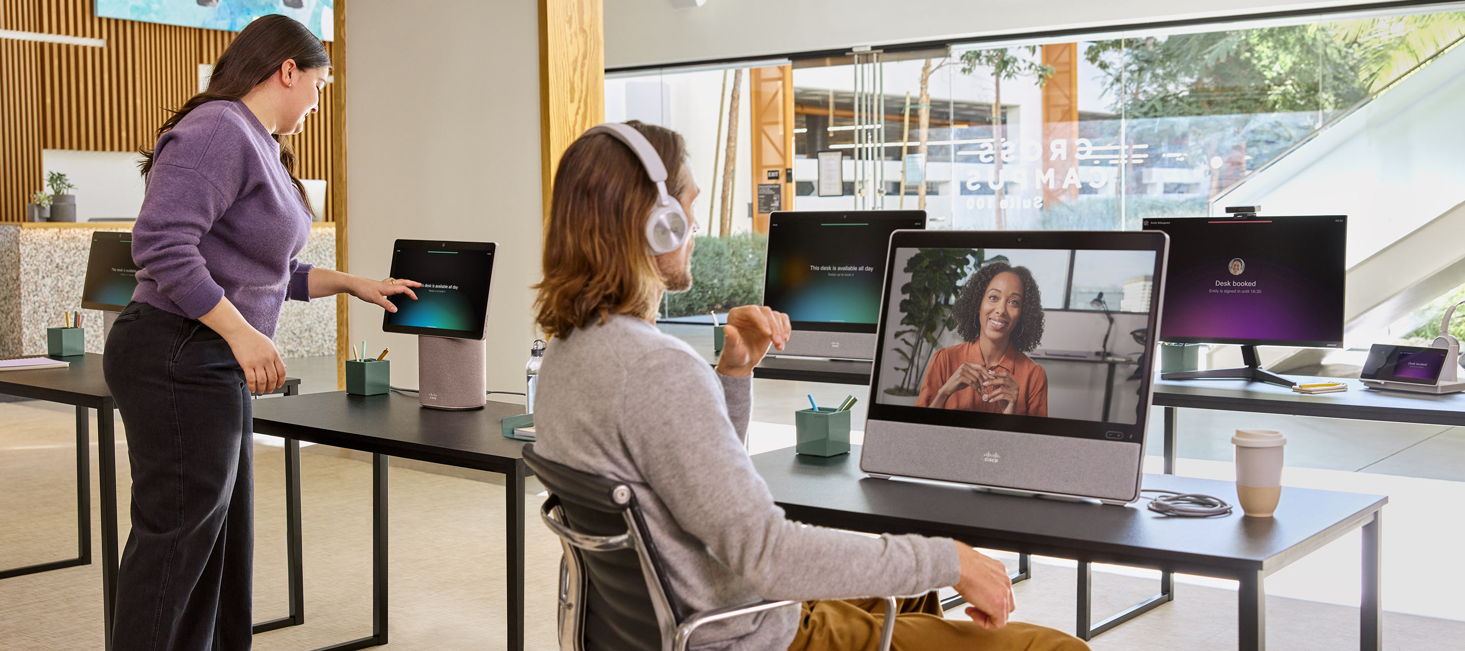 Sitting at a desk in an open office, a professional wearing a headset video conferences with a colleague.