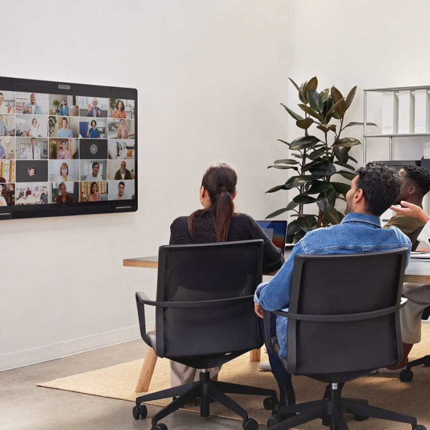 A video conferencing meeting. Three in-office employees view a mounted screen showing their teammates.