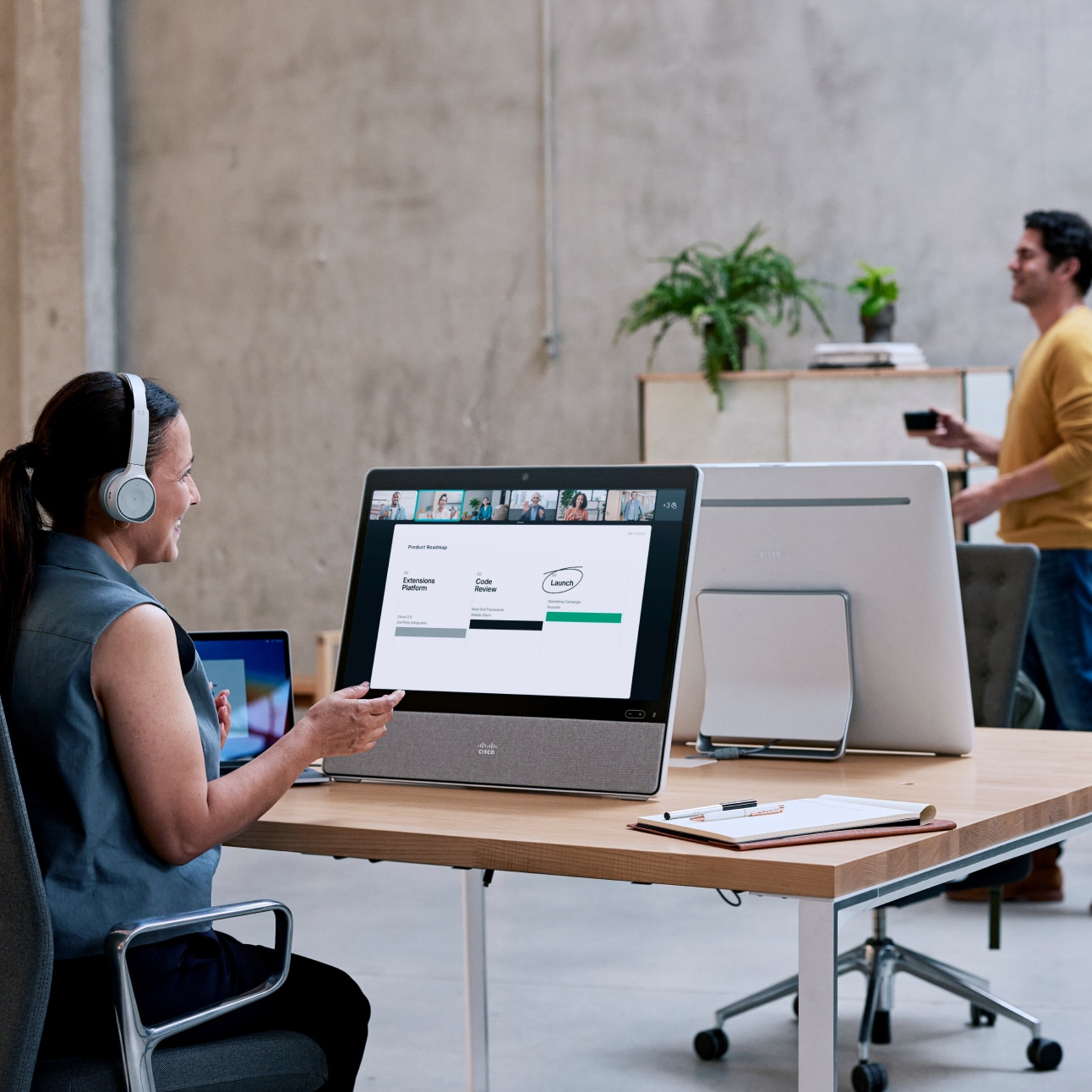 Sitting at a desk in an open office, a professional wearing a headset video conferences with colleagues via a Webex Desk.