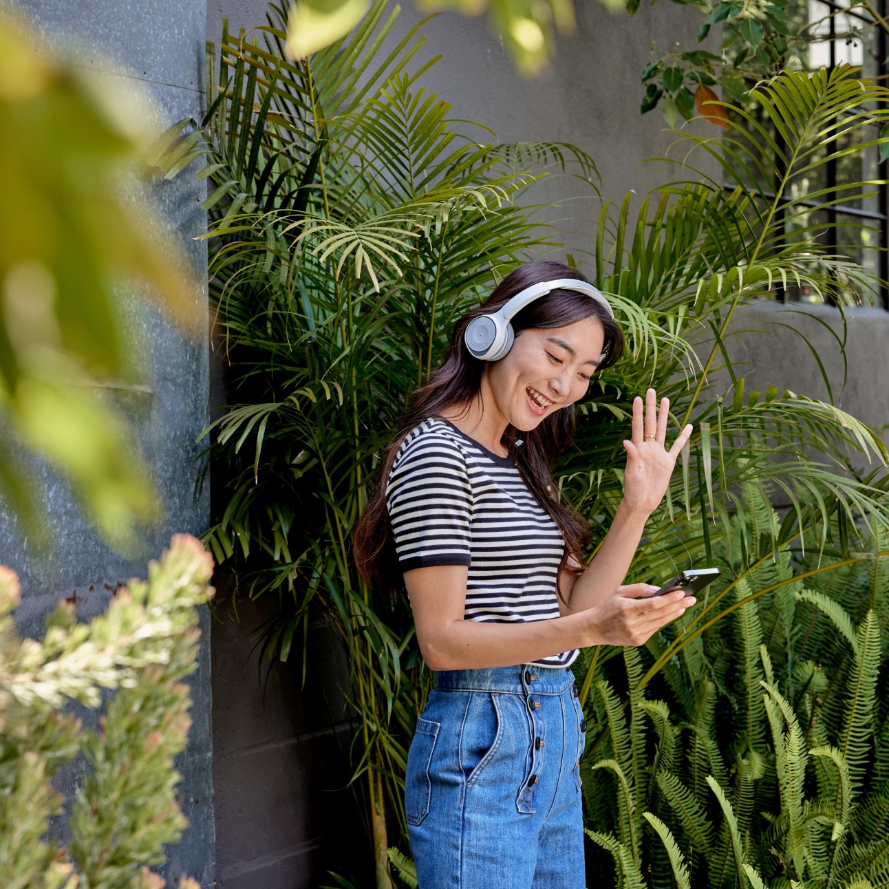 Standing in front of a gray building by green foliage, a person in a headset collaborates on a mobile phone via Webex Calling.