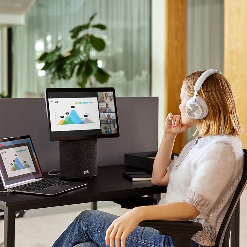 Woman at her desk in the office watching a presentation during a video conference on a Cisco Desk Mini while wearing a Bang & Olufsen Cisco 980 headset.