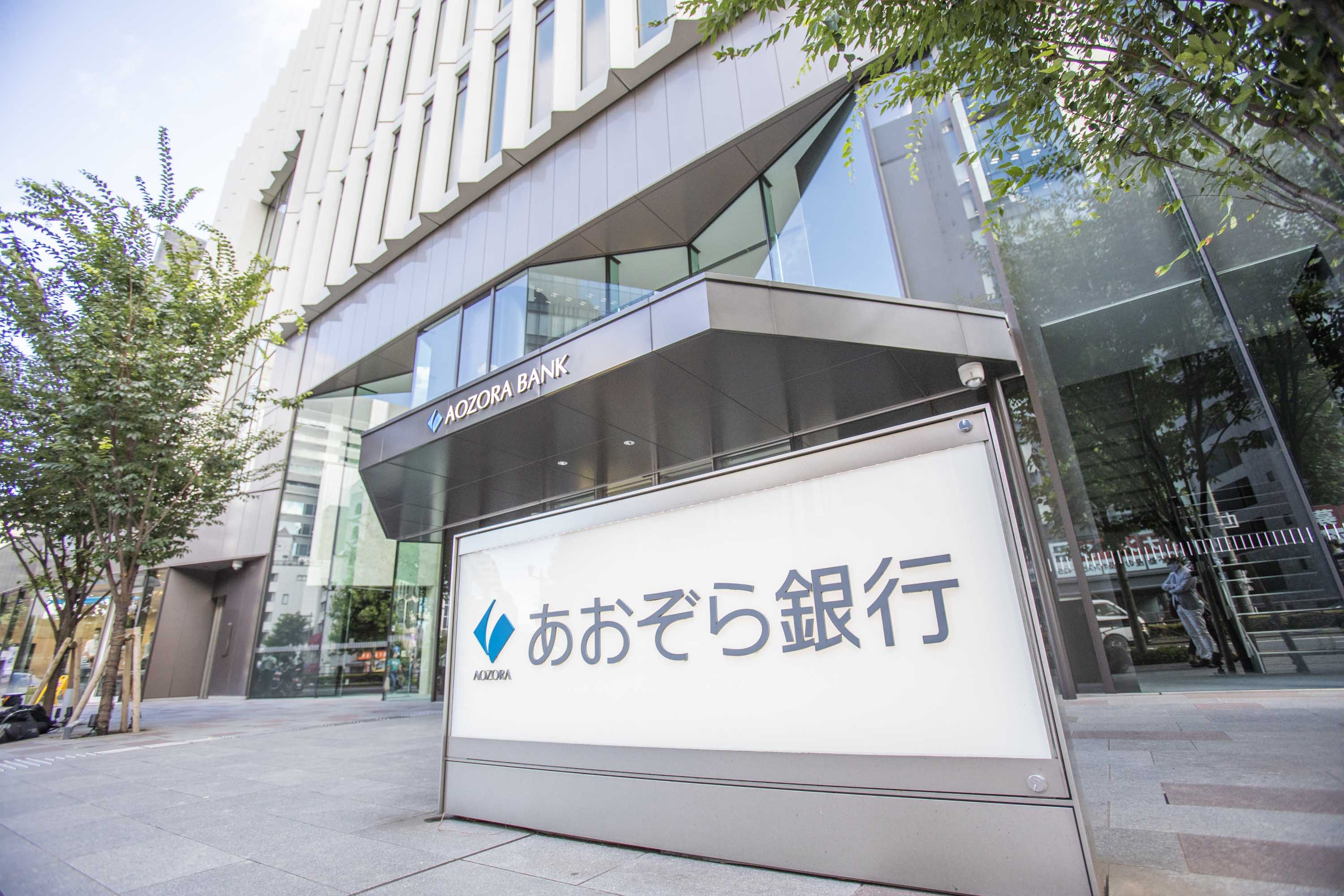 The exterior of the Aozora Bank office on a tree-lined street. The name Aozora Bank is visible above the glass office doors.