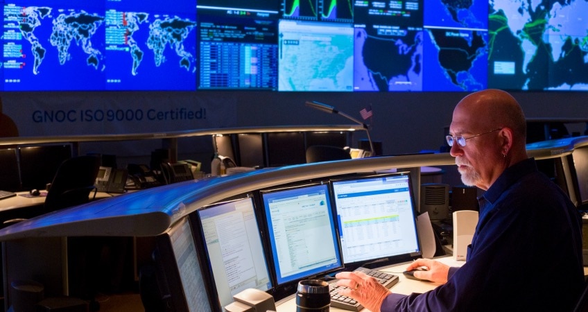 A man sits at a desk looking at four monitors. Large screens line the room walls in front of him, showing maps and charts.