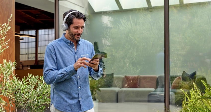 Person in headset and blue shirt collaborates outside on his mobile phone. He stands near some plants and a big window.