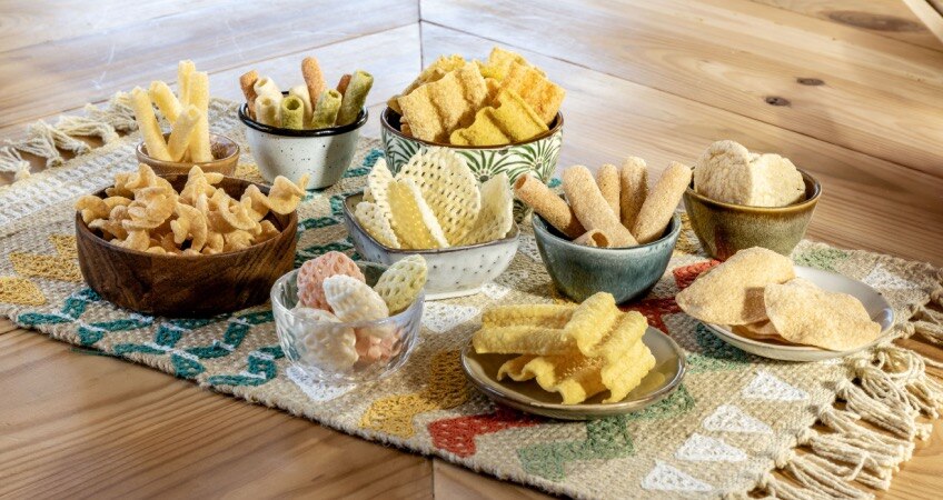 Several types of chips and snacks on little plates and bowls are displayed on a colorful woven placemat atop a wooden table.