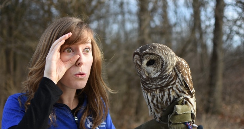 Person stands outdoors in front of several trees, holding an owl. She widens her own eyes, appearing to imitate the owl.