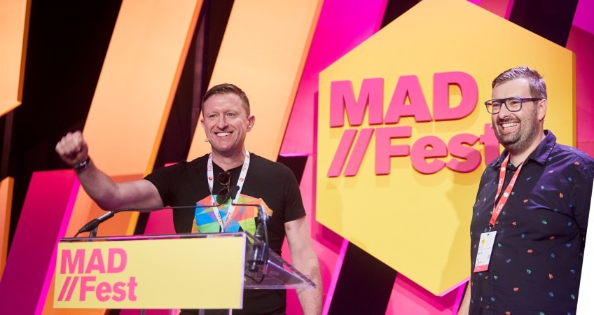 Two people smile onstage at a MAD//Fest event. The decor is bright pink, yellow, and orange.