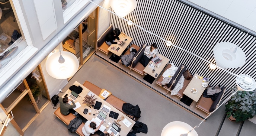 People work in a modern open workspace with high ceilings, lots of light, 3 smaller tables, and a long table in the center.