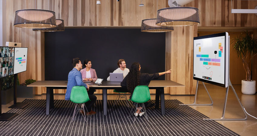 Four colleagues collaborate in a conference room while others video conference in, looking at content on a Webex Board 85S.