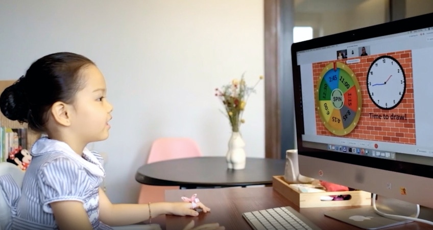A young child sits at a desk, looking at a computer screen, engaged in virtual learning via video conferencing.
