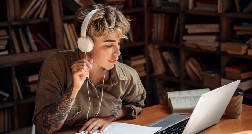 Sitting at a desk, a student in a headset engages in online learning on a laptop, surrounded by books and a coffee cup.