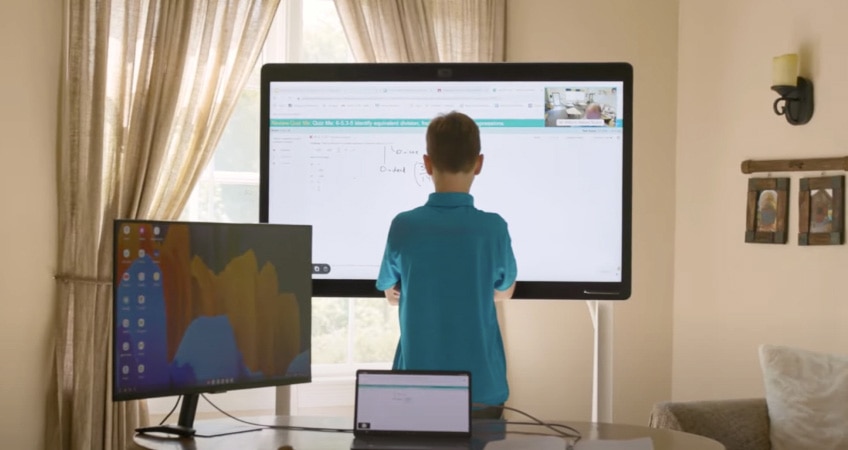 A young boy stands in front of a Webex Board in his home, engaged in virtual learning.