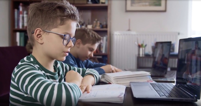 Two young children sit side-by-side at a desk, each engaged in virtual learning on their own laptops.