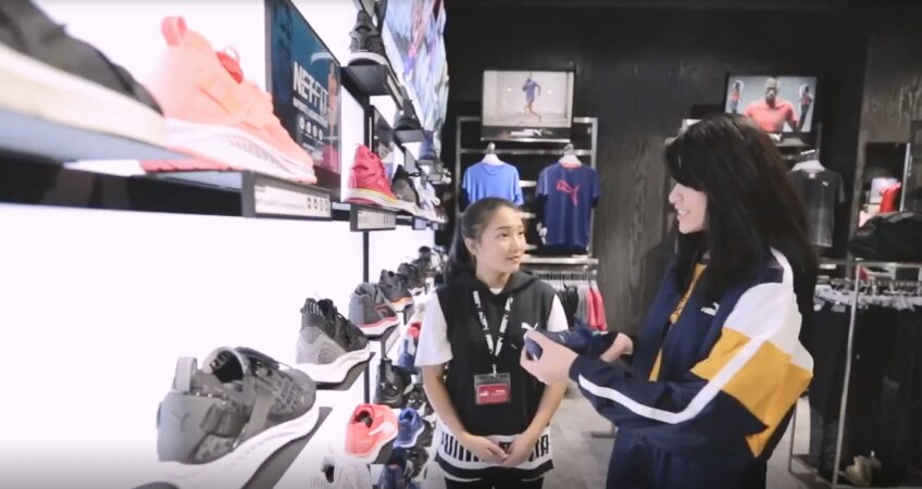 Two people chat in a PUMA store near shelves with many pair of shoes displayed on them.