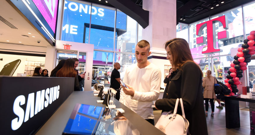 Two people look at a mobile phone in a large, bustling room with a giant pink T-Mobile logo on the wall behind them.