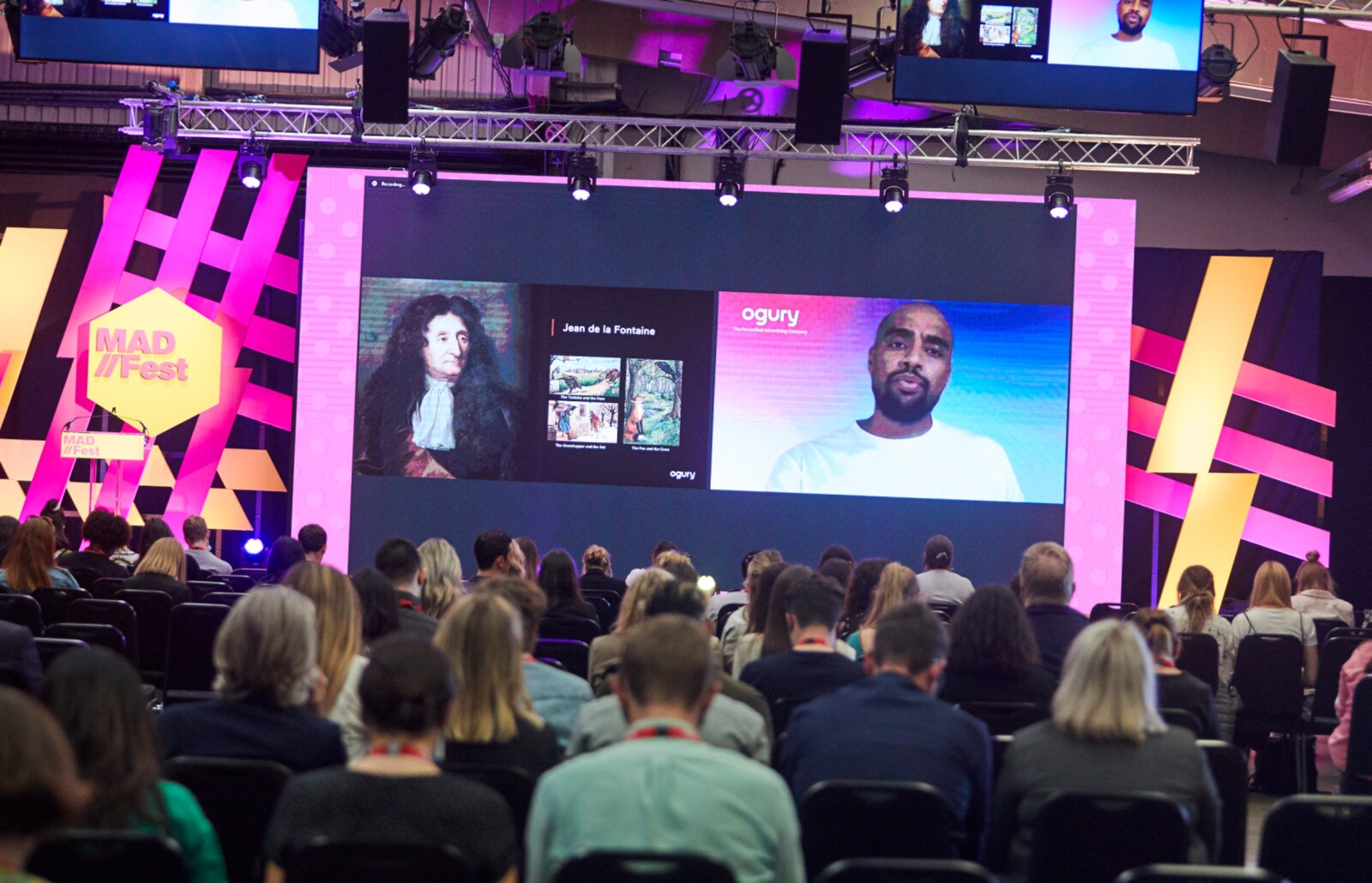 In-person attendees listen while a virtual presenter speaks on the big screen at the MAD//Fest event.