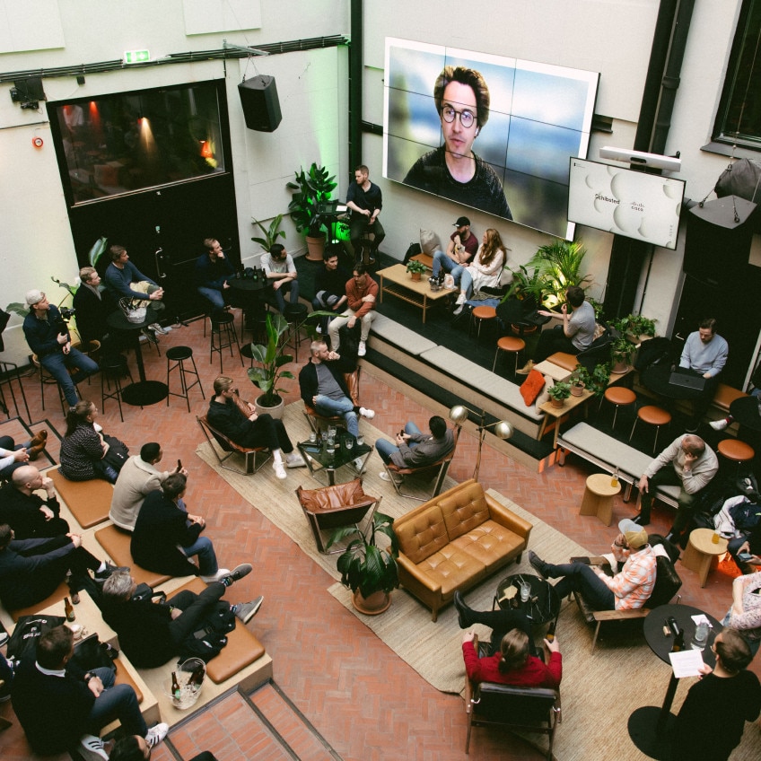 People in an open meeting area listen to a presentation via video meeting. The presenter is seen on huge screen on one wall.