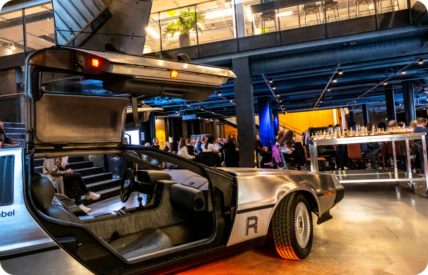 Event in progress at a Rebel space. A silver car is on display in the foreground. A large audience sits in the background.
