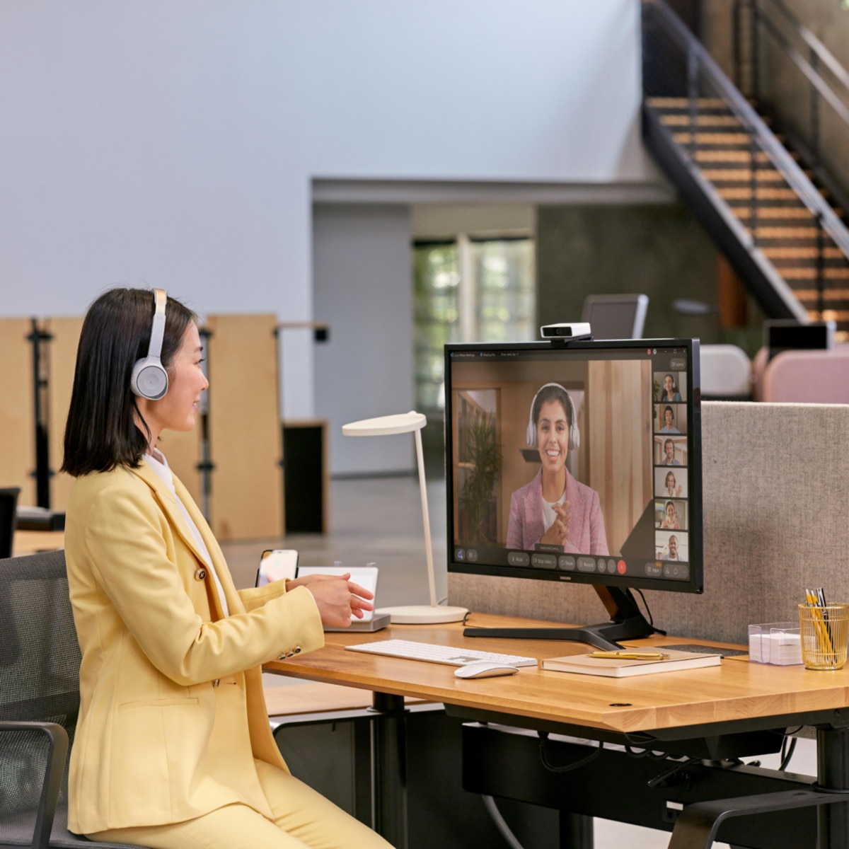A professional in a pale yellow suit and a headset sits at a desk in an office, video conferencing with several colleagues.