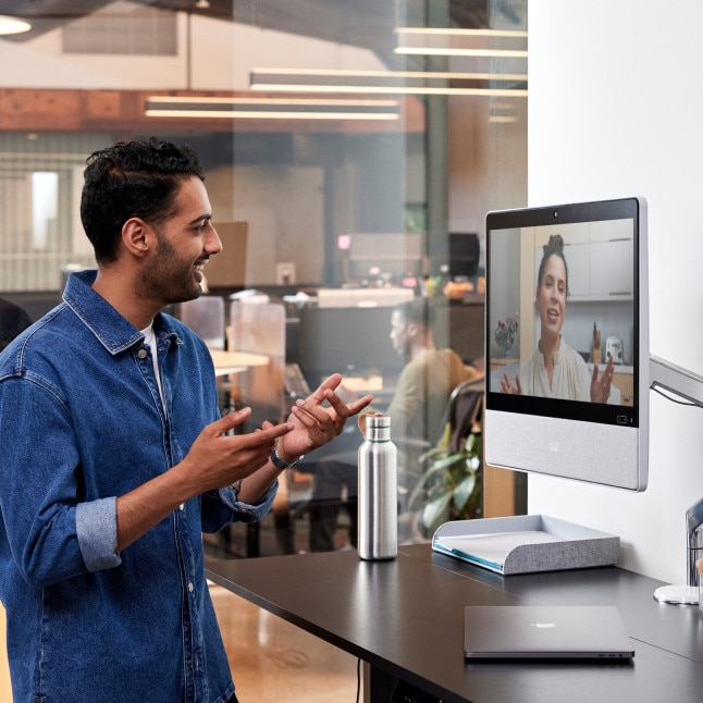 Person in a denim button down shirt at a standing desk in an office collaborates with a colleague via a video call.