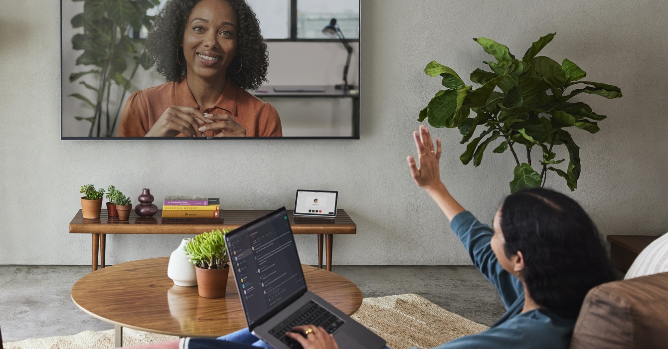 A person gives a friendly wave to a person on a larger monitor.