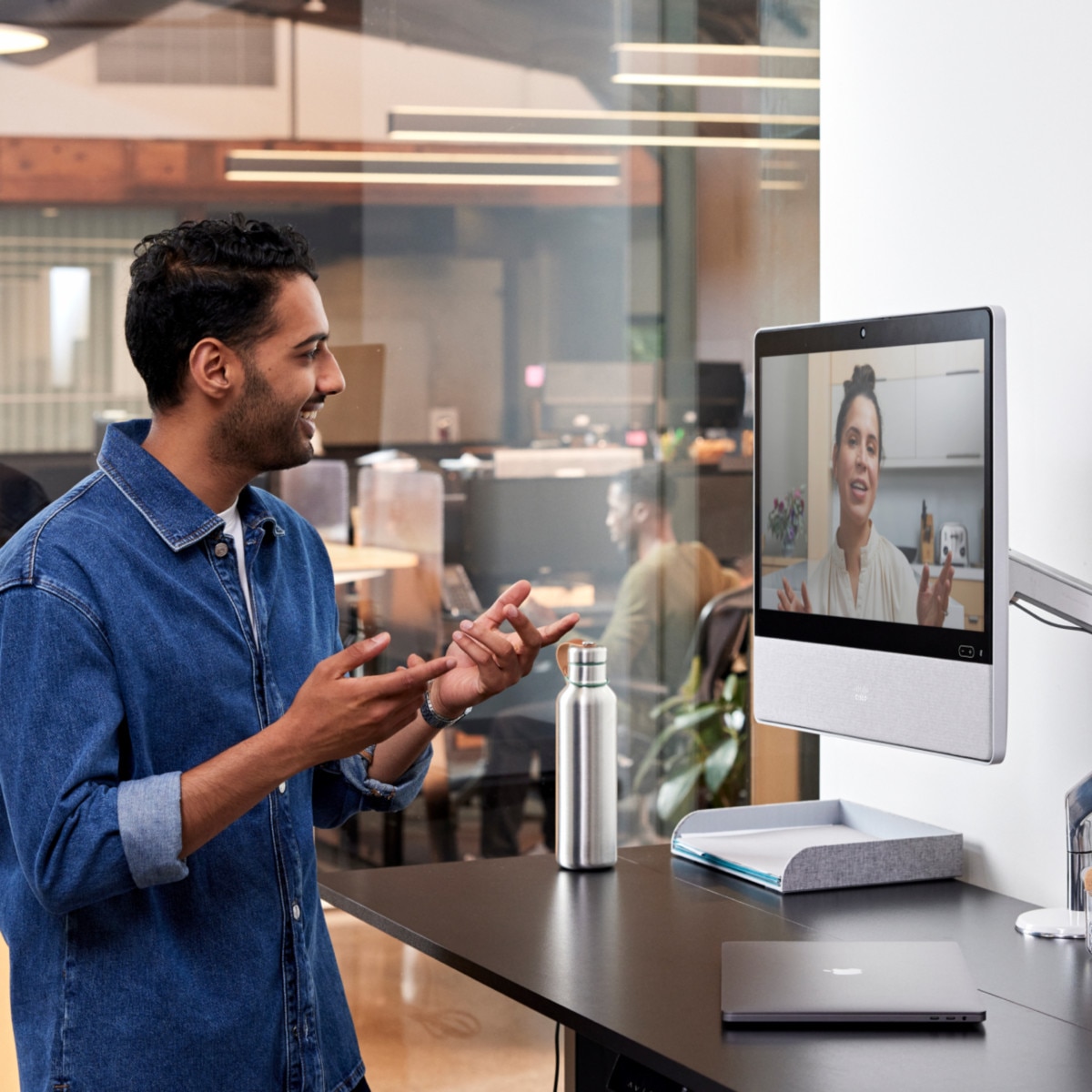 Person in a denim button down shirt at a standing desk in an office collaborates with a colleague via a video meeting.