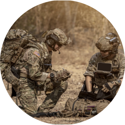 A soldier in combat gear kneels in a field and consults a tablet while another soldier looks on.