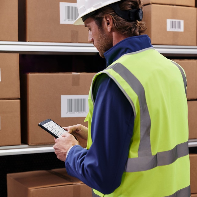 A frontline worker in a safety helmet and reflective vest consults a mobile phone while standing near shelves of cardboard boxes.