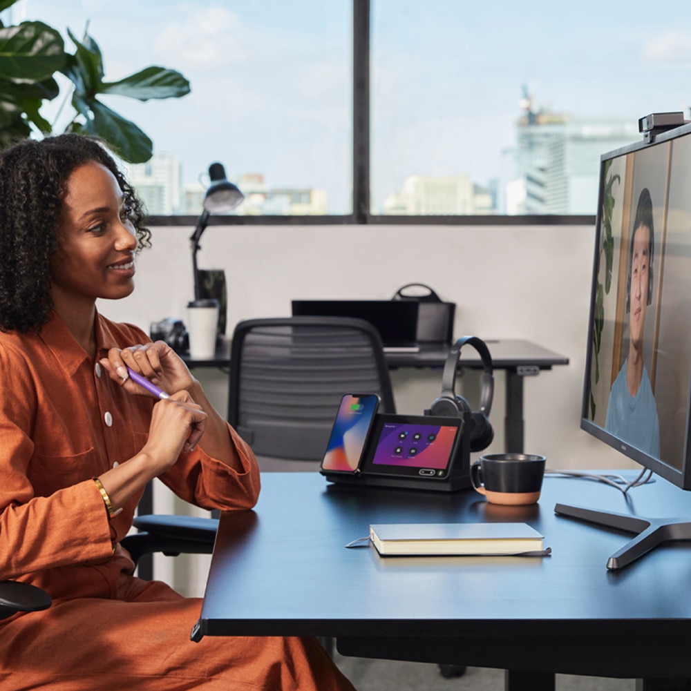 A person smiles while sitting at a desk with a notebook, cup, and a Webex Desk Hub.