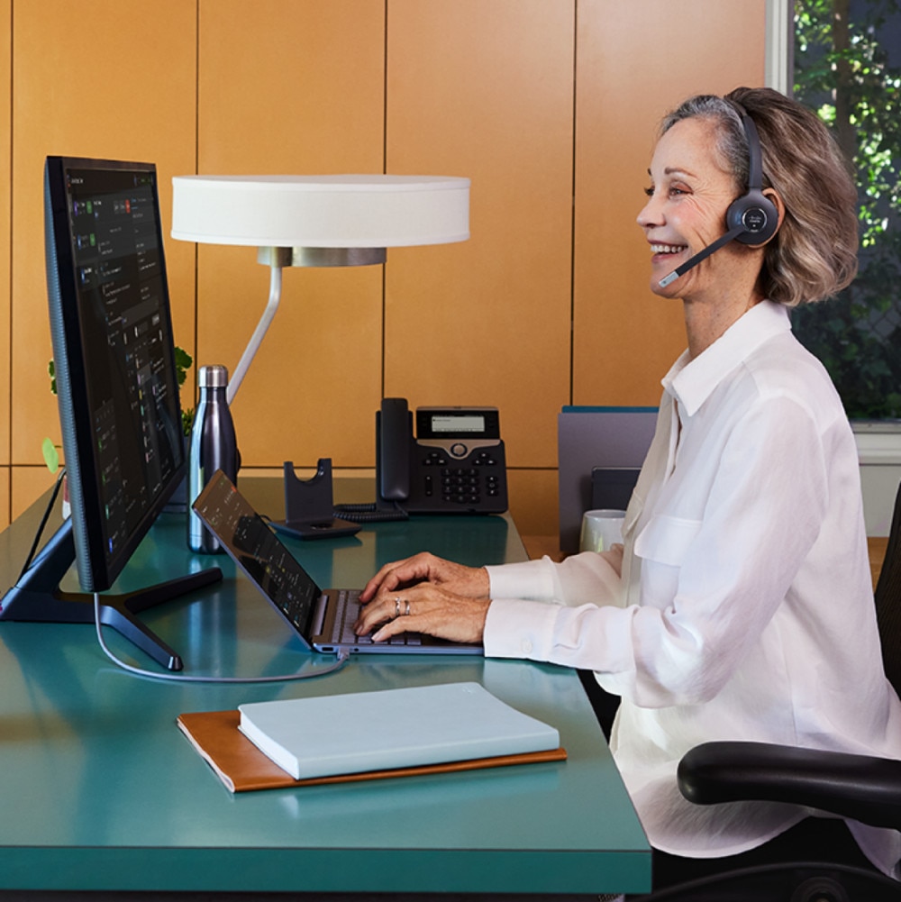 A customer service agent smiles while typing on a laptop and wearing a headset.