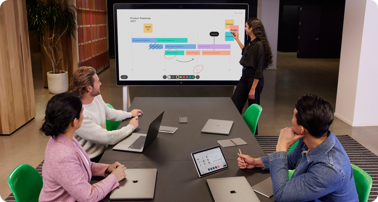 Three colleagues sit at a table in a meeting room, listening as one colleague talks at the front. Everyone has MacBooks.
