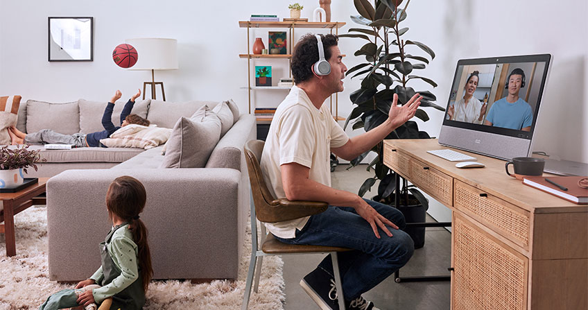 Remote worker speaks in virtual meeting from living room using Cisco Desk Pro while children play behind him.