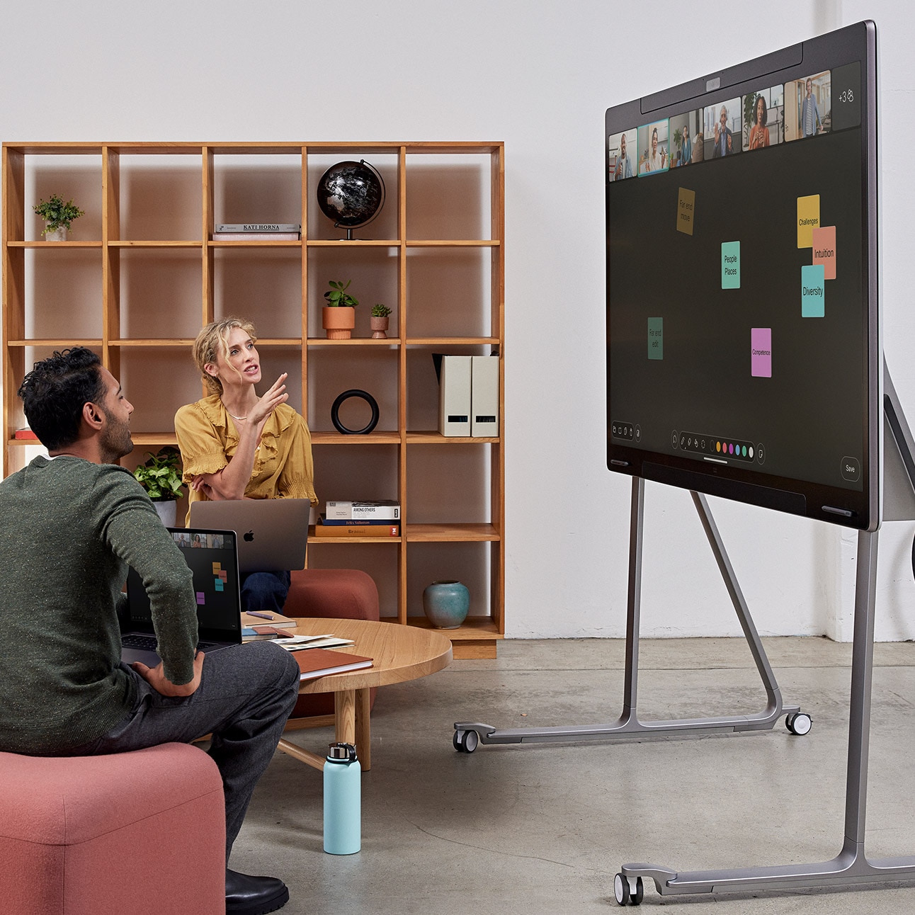 Two people collaborating in an open huddle space with the Cisco Board Pro on wheel stand. The touch board is used for digital whiteboarding with digital sticky notes shown on the canvas and the live video of remote participants on top of the ideation area.