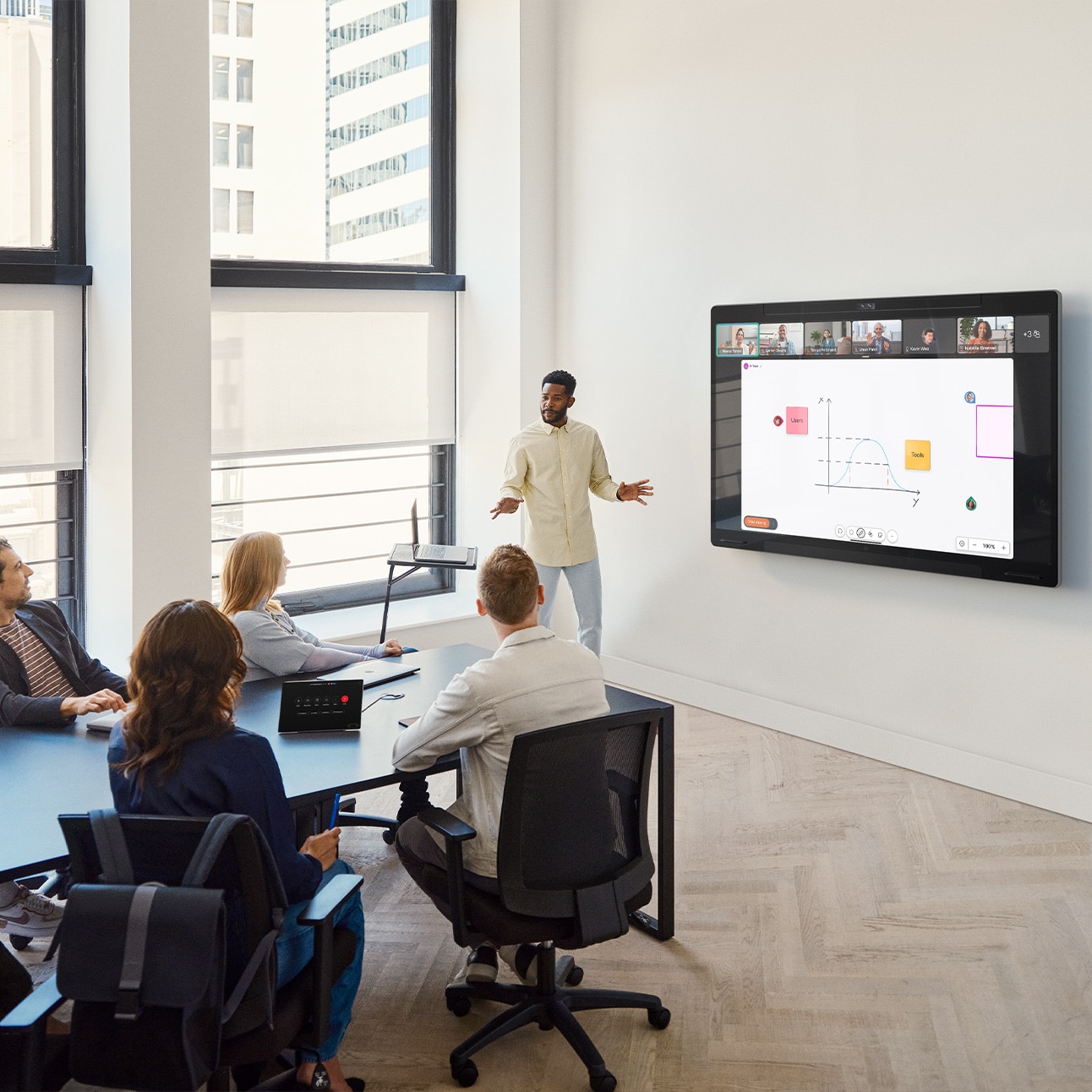 Five people collaborating in a medium-sized conference room using the wall-mounted Cisco Board Pro and the tabletop Cisco Room Navigator control panel. One person is standing close to the Board Pro to hold a presentation using the touch screen which shows a digital whiteboard canvas with sticky notes, shapes, and the live video of remote video meeting participants.