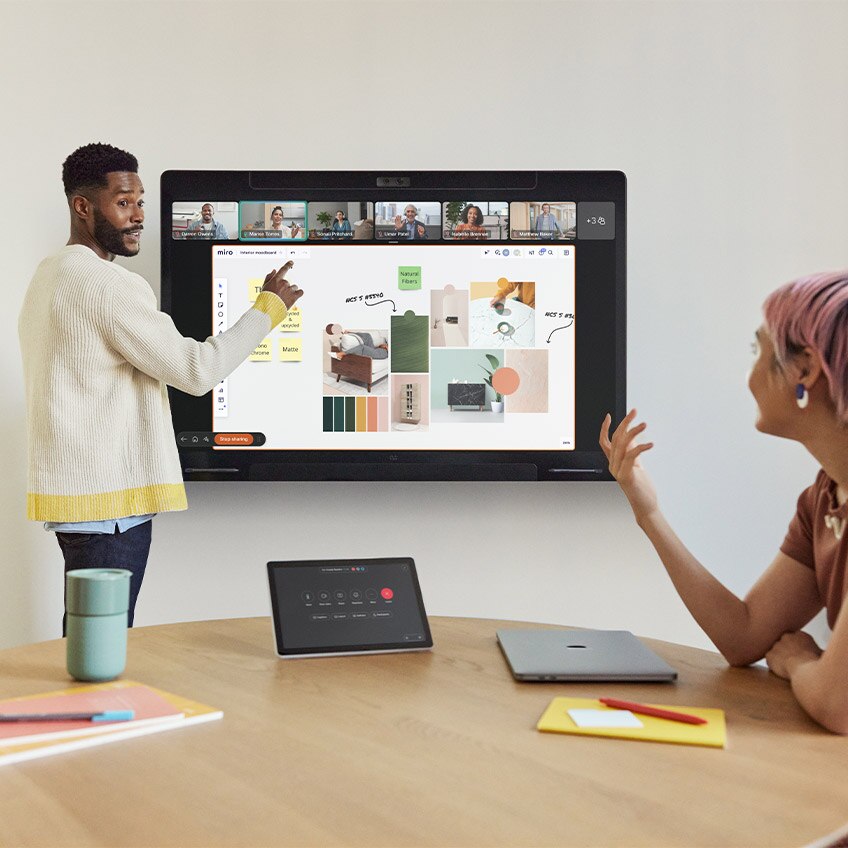 Two people attending a design sync in a small huddle room using the wall-mounted Cisco Board Pro and the tabletop Cisco Room Navigator control panel. One person is standing in front of the Board Pro to draw and annotate on the touch screen which shows an active web application with renders of product prototypes.