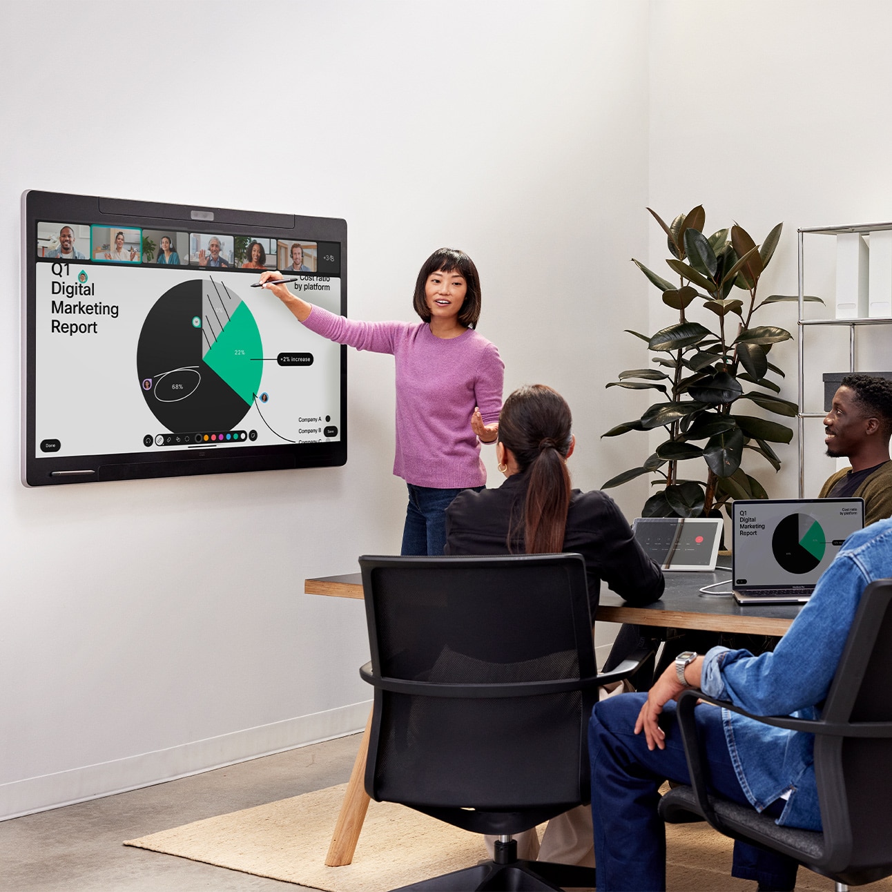 Four people collaborating in a small meeting room using the wall-mounted Cisco Board Pro as a room conferencing device, the tabletop Cisco Room Navigator control panel and a laptop on the tabletop. One person is standing close to the Board Pro and is using a board pen to annotate on the shared content that shows a pie chart, notes, arrows and the live video of remote video meeting participants.