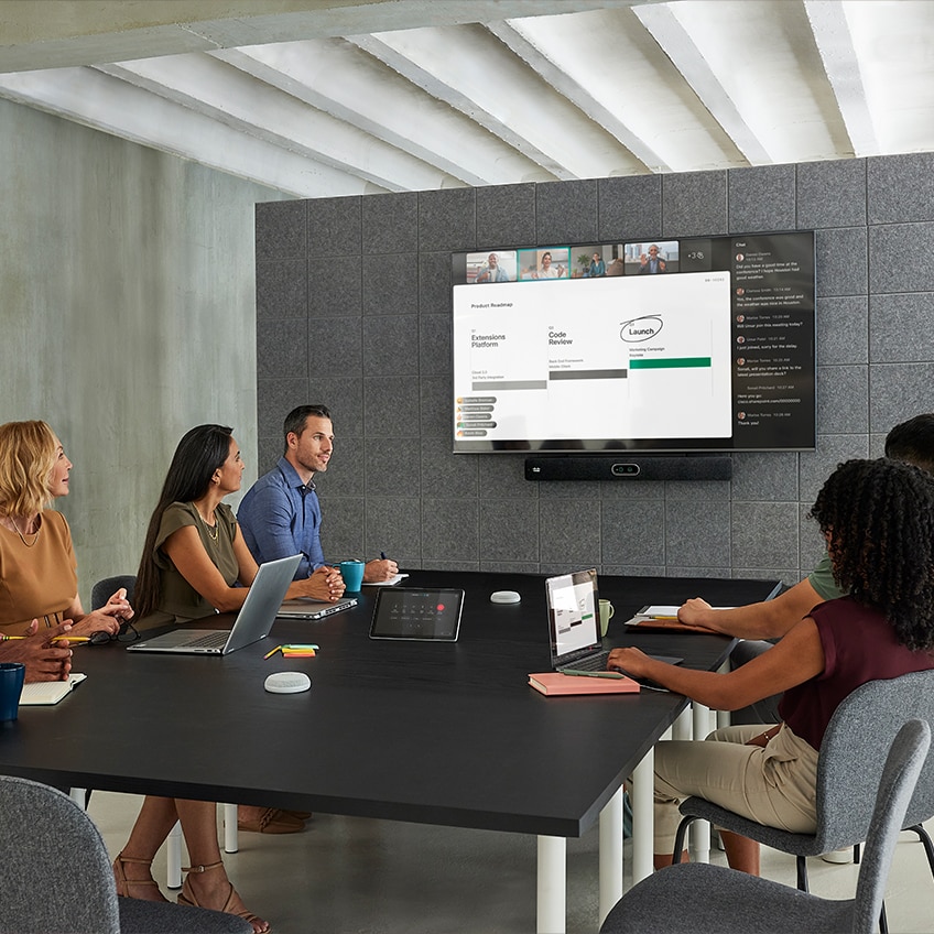 Five professional sitting at a meeting room table in a video conferencing meeting.  The Cisco Room Bar Pro is mounted to the bottom of the display at the front of the room.