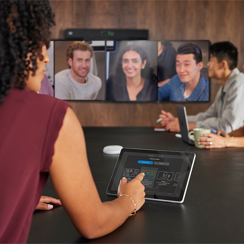 A professional interacts with the Cisco Room Navigator on a meeting room table while in a video conference using the Cisco Room Bar Pro video bar.