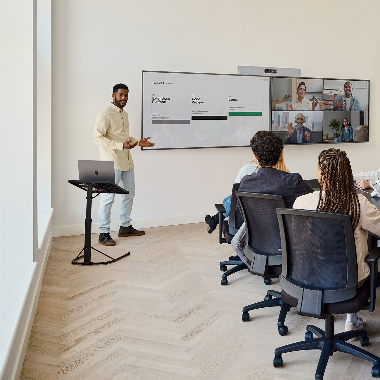 Five people collaborating in a meeting room with a man standing at the front of the room. Room Kit EQ mounted on top of two displays showing remote participants and a presentation.