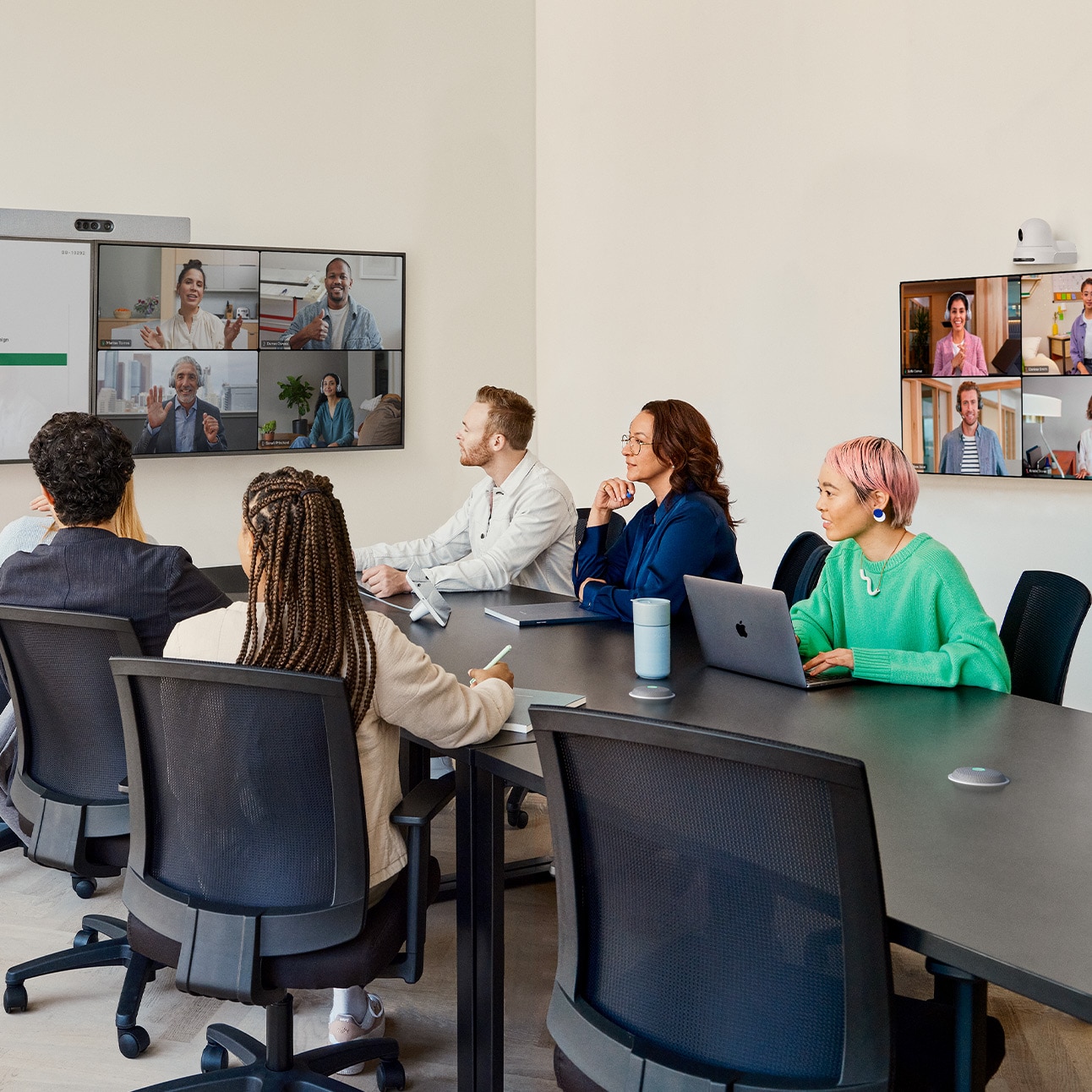 Six people collaborating in a conference room. The Cisco Quad Camera bar is mounted on the wall above dual external displays showing shared content and remote participants of a live video meeting. The Cisco PTZ 4K Camera is mounted on the sidewall above an external display showing remote meeting participants. The team is using collaboration peripherals including the tabletop Cisco Room Navigator room controller and two Cisco Table Microphone Pro devices placed on top of the table surface.
