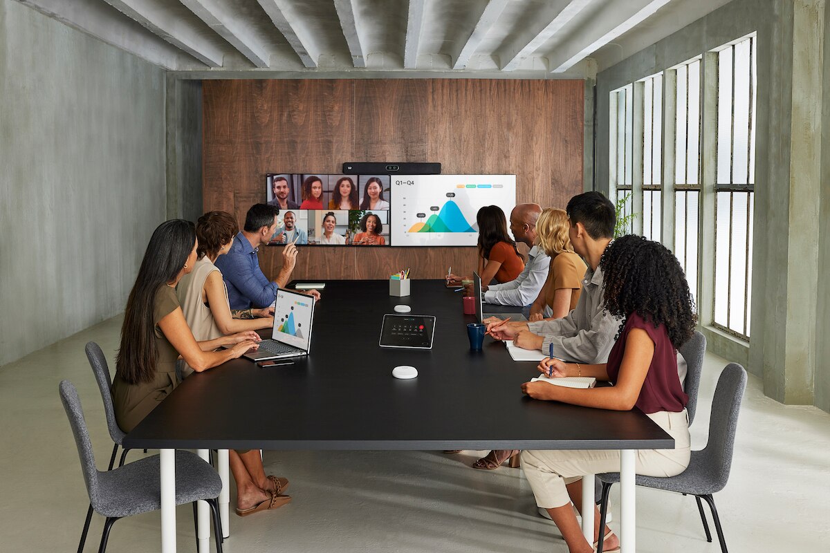 A large executive meeting room showing the integrated, triple-screen Cisco Room Panorama video conferencing system with Cisco wall finish and displaying the Cisco RoomOS home user interface, alongside the tabletop Room Navigator room controller, and Cisco Table Microphone Pro units on the conference table.