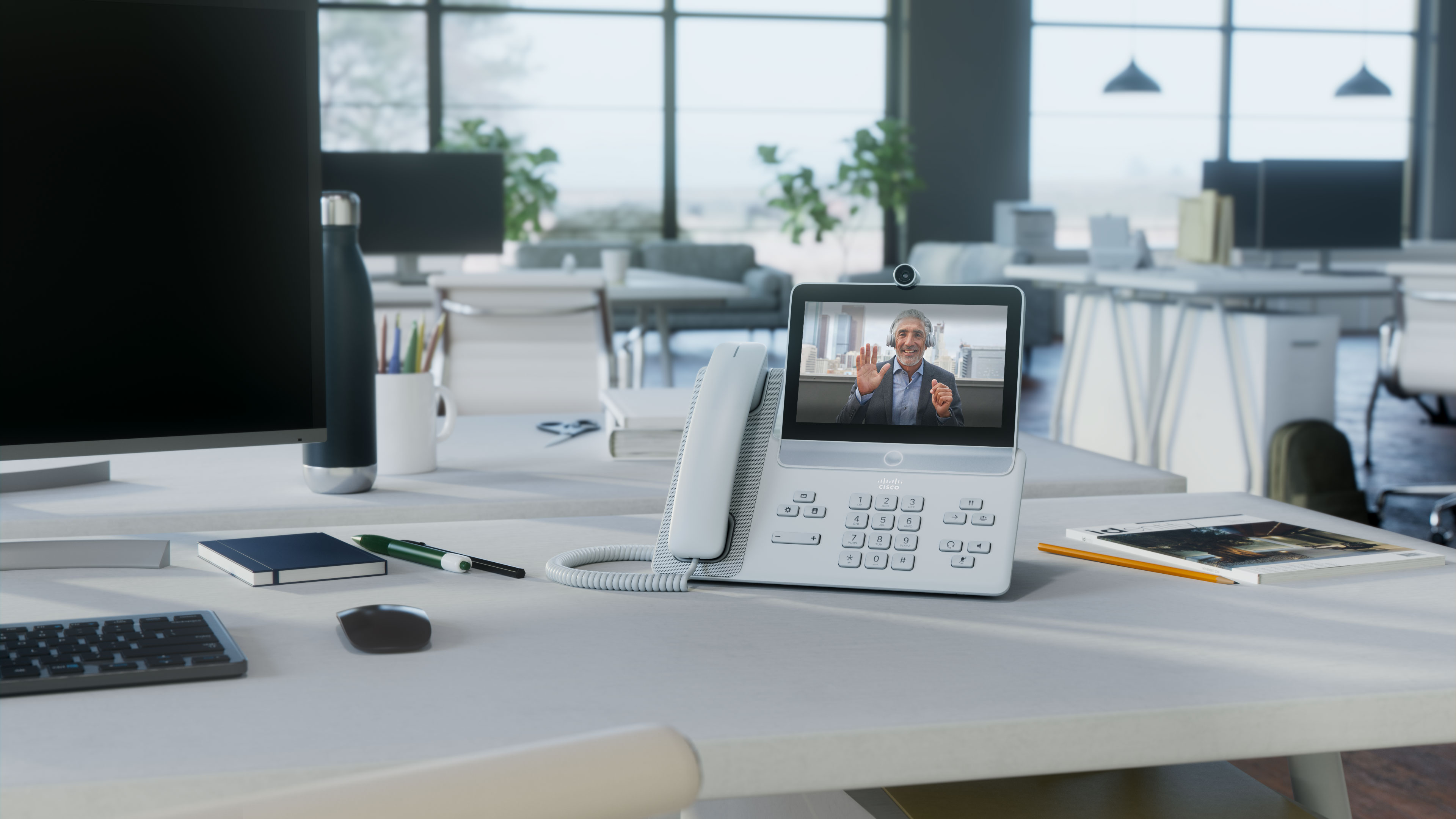 The Cisco Video Phone 8875 on a desk at a workspace in a sunny open office.