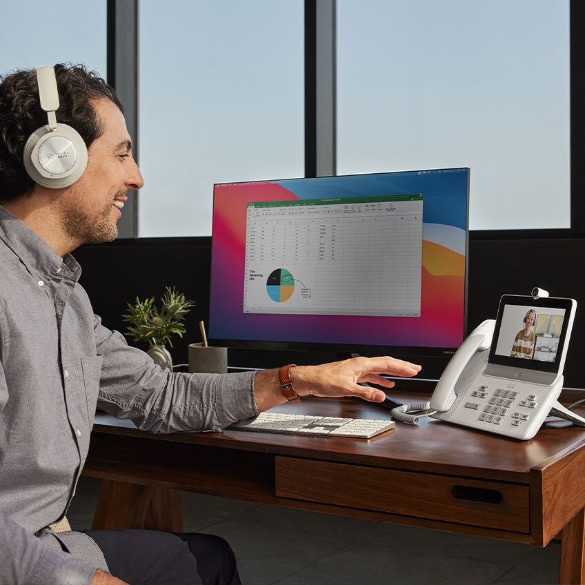 Person in a headset video conferences using the Cisco Video Phone 8875. His colleague is visible on the phone's screen.