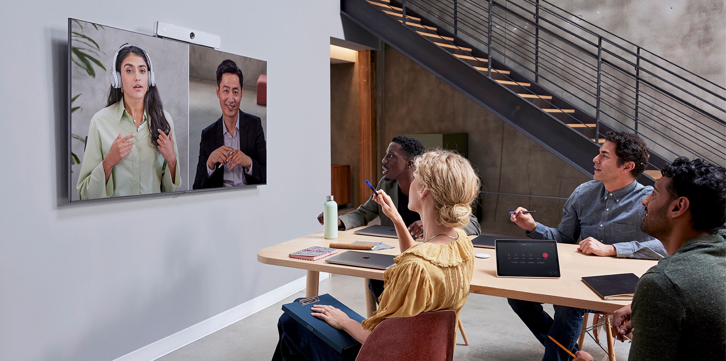 4 colleagues in an open shared space collaborate with 2 colleagues via a video meeting using the Cisco Room Bar video bar.