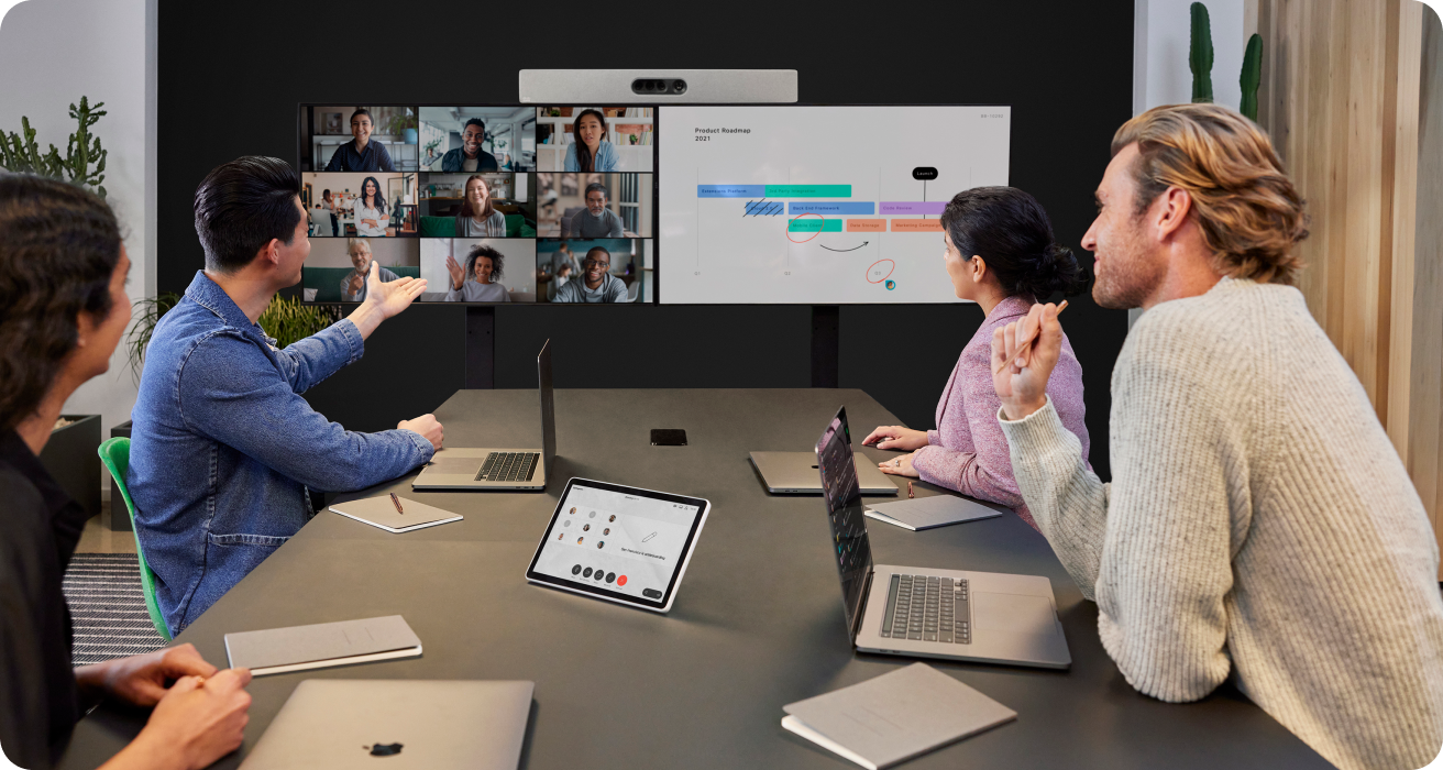 Four colleagues collaborate in a conference room with nine other colleagues who have video conferenced in.