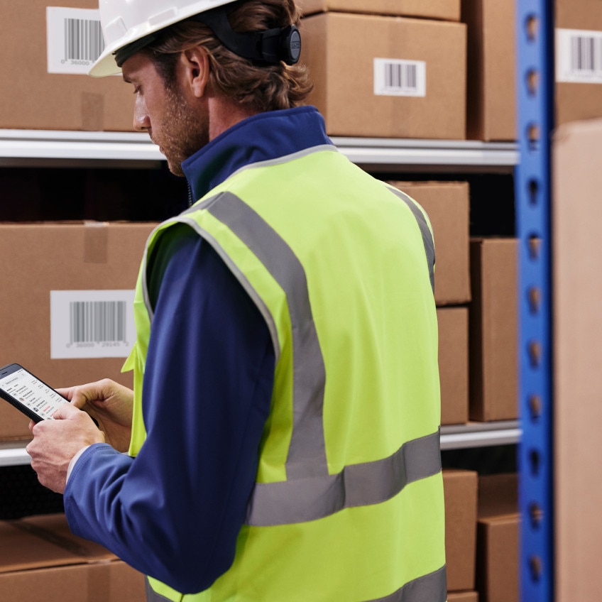 An employee in a hard hat and safety vest works on a smartphone near a shelf of boxes.