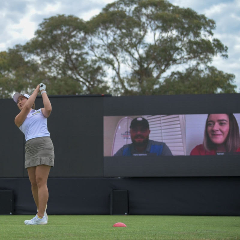 Golfer swings at a Webex Player Series tournament, while remote attendees look on from a screen near the green.