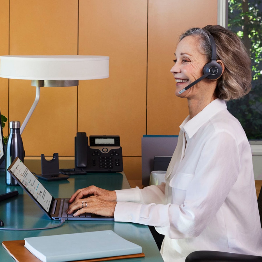 A professional wearing a headset smiles while typing on a laptop.