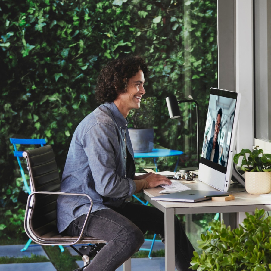 A person is seated at a desk, smiling at a person on the monitor.